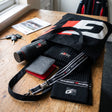 A black tote bag, matching black knit beanie, black wrist strap, and black wallet with red lining, all branded with the GAZOO Racing logo, arranged on a wooden table next to keys and a magazine. A black and red racing jacket is visible hanging in the background.